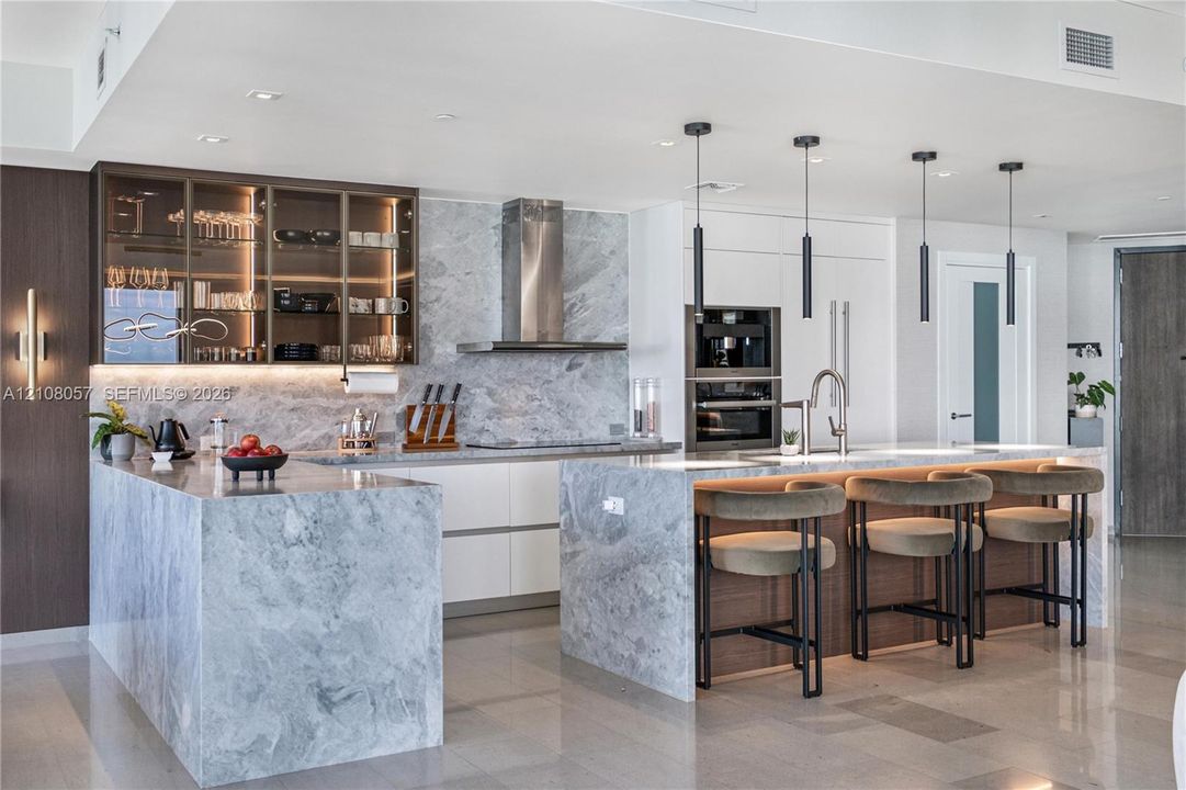 Kitchen featuring stone-clad islands and countertops, integrated stainless steel appliances, upper cabinetry with glass-front display panels, matte black pendant lighting, and flat-panel cabinetry