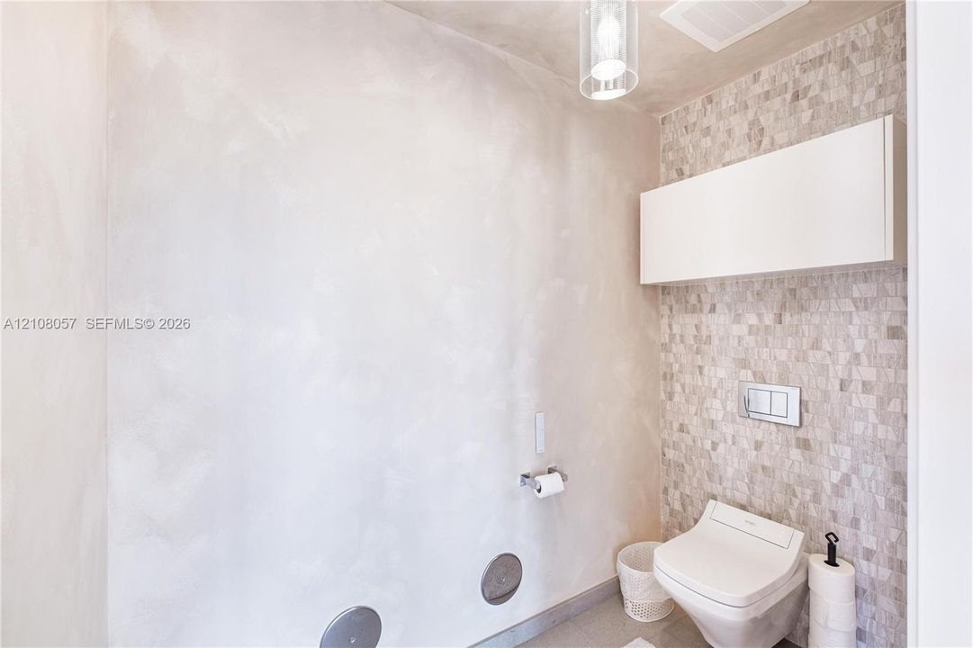 Wall-mounted toilet with a dual-flush actuator plate, mosaic tile accent wall, wall-mounted storage cabinet, and glass pendant lighting
