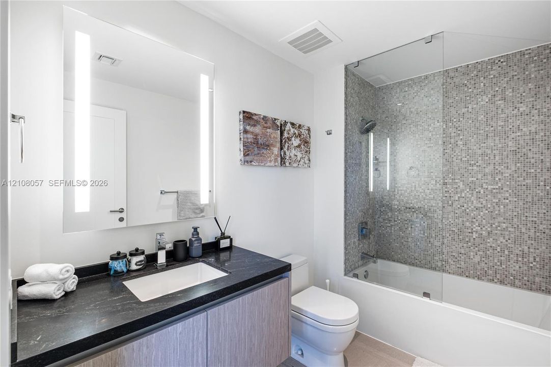 Vanity with a dark countertop and integrated rectangular sink, wood-finish cabinetry, illuminated mirror, and a tiled shower-tub combination with a glass panel
