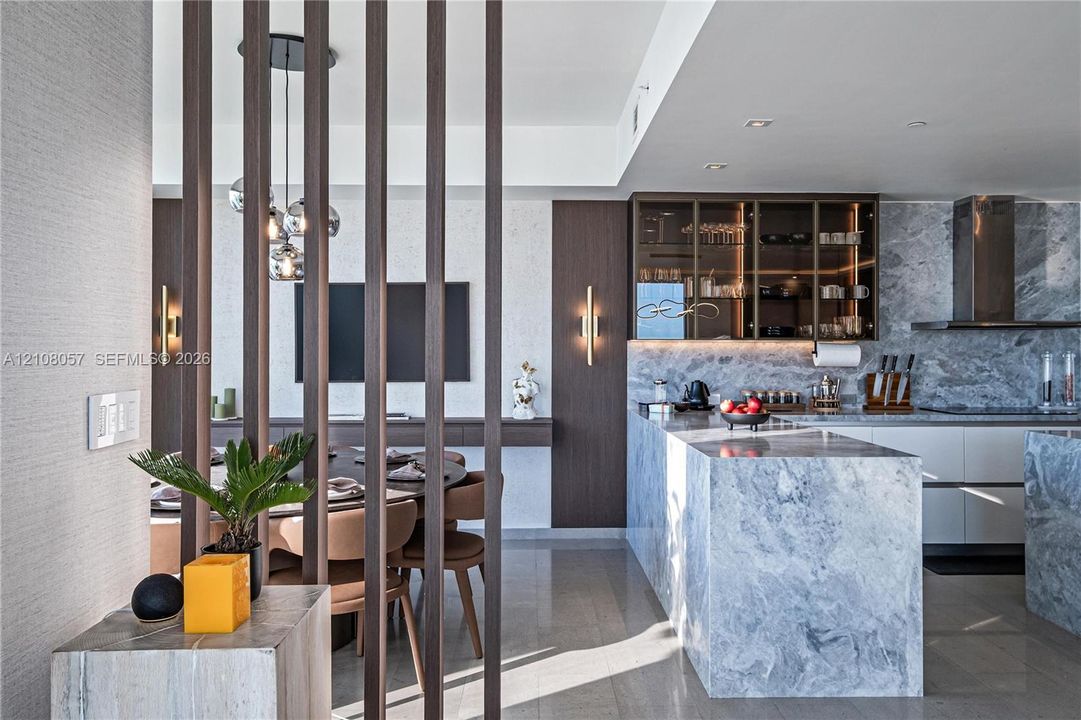 Kitchen featuring stone-clad waterfall island and backsplash, custom cabinetry with glass-front uppers, decorative vertical wood slat room divider, and integrated wall-mounted lighting