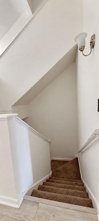 Staircase featuring neutral-toned carpeting, white painted walls, a wall-mounted light fixture with a frosted glass shade, and a handrail