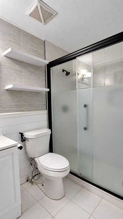 Shower enclosure with sliding frosted glass doors, matte black hardware, wall-mounted white shelving, horizontal board wall paneling, and tile flooring
