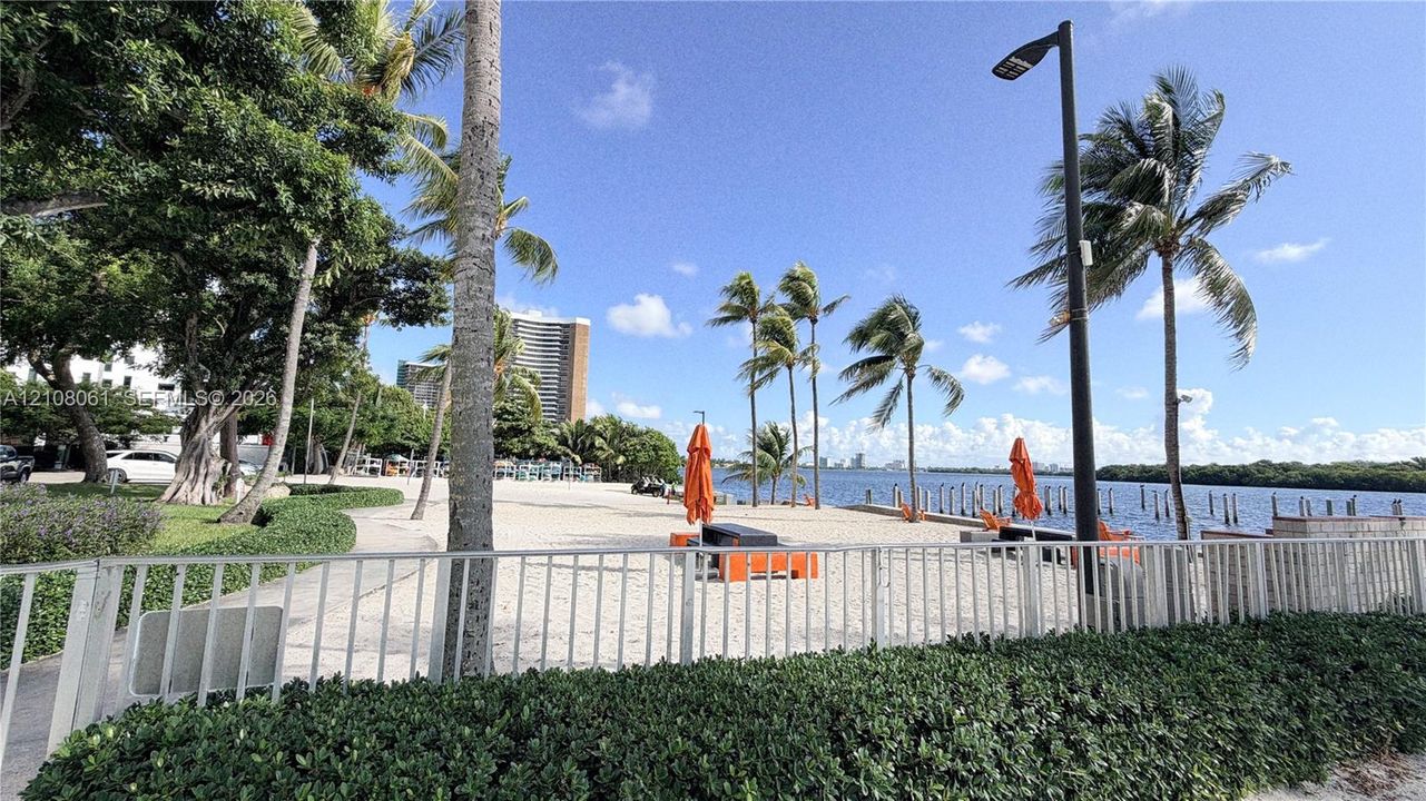 Waterfront property featuring a sandy shoreline, mature palm trees, professional landscaping, and metal perimeter fencing