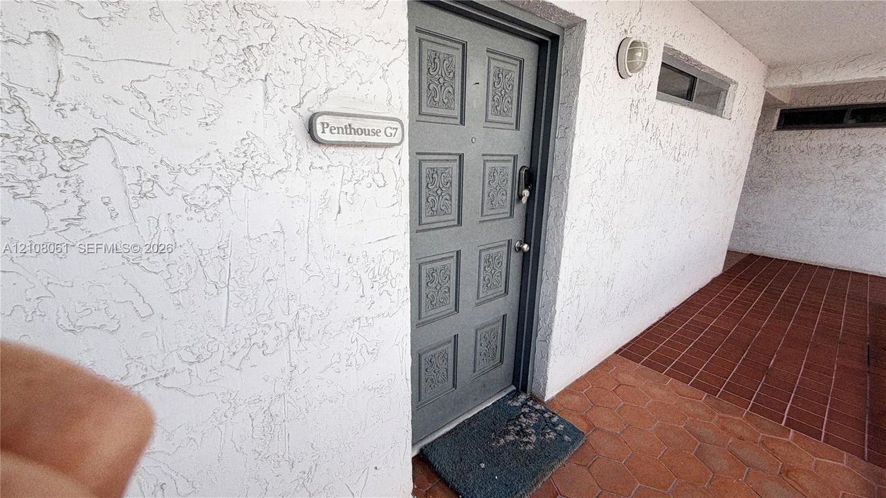 Gray-toned door with ornate paneled detailing, textured stucco exterior wall, and red hexagonal tile flooring