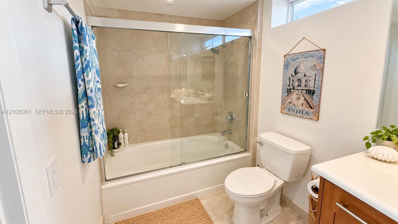 Bathtub with a glass sliding enclosure, tile wall surround, built-in soap dish, and a wood-finish vanity with a white countertop
