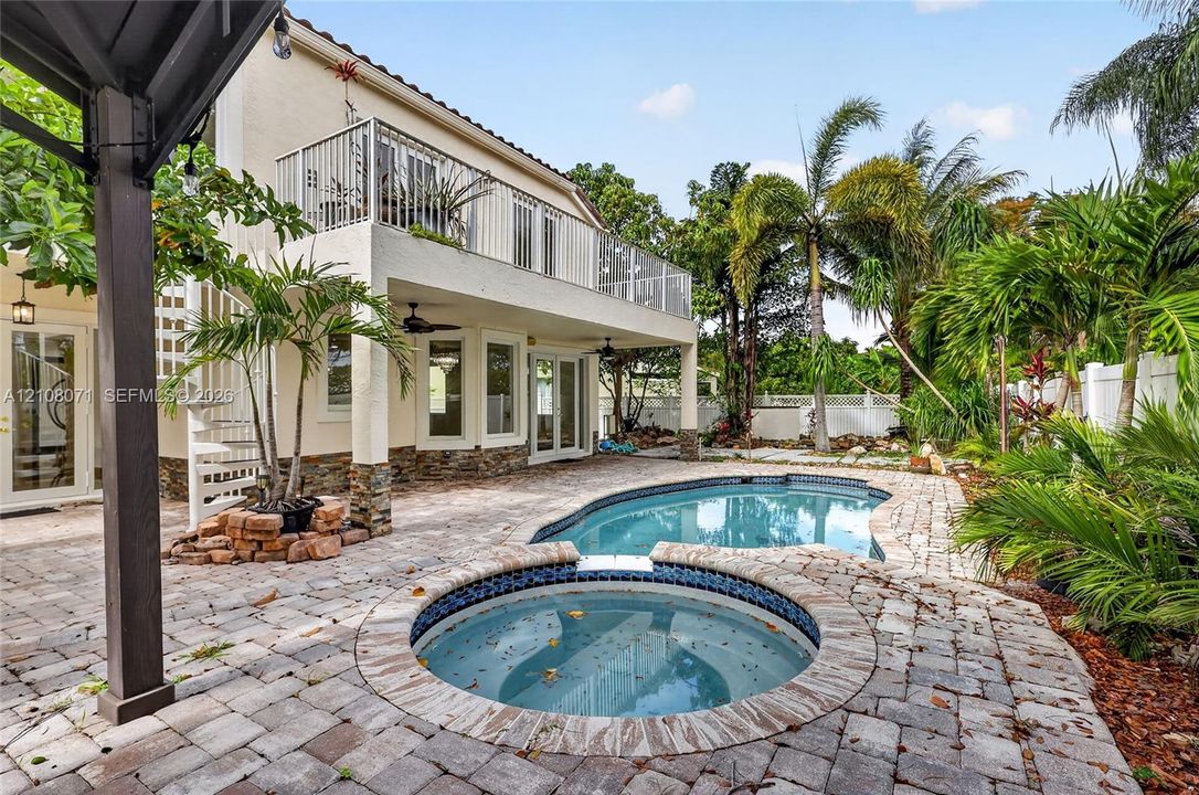 Stucco exterior with stone veneer detailing featuring a second-story metal balcony, spiral staircase, in-ground pool, and integrated circular spa surrounded by paver hardscaping