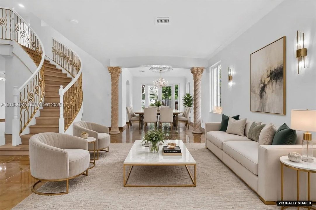 Curved staircase with ornate gold-tone metal railing, decorative stone columns, polished stone flooring, and multi-tiered crystal chandelier