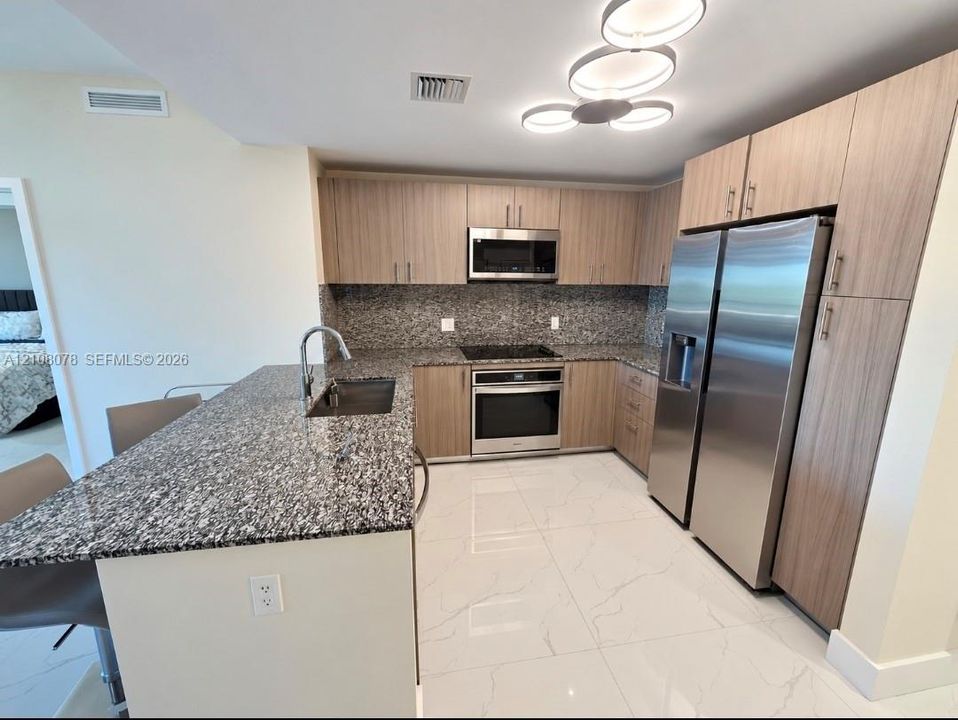 Kitchen featuring flat-panel wood-finish cabinetry, speckled stone countertops, large-format tile flooring, stainless steel appliances, and a modern multi-pendant light fixture
