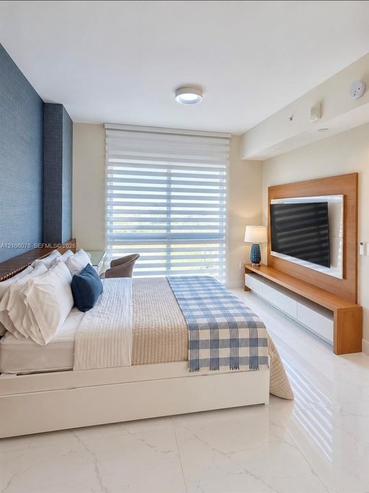 Neutral-toned bedroom featuring polished tile flooring, a blue textured accent wall, integrated wood-finish media shelving, and dual-layered window shades