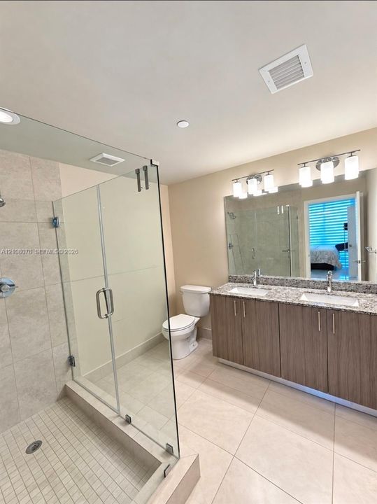 Bathroom featuring a dual-sink vanity with stone-finish countertops, frameless glass shower enclosure, large wall-mounted vanity mirror, and neutral tile flooring