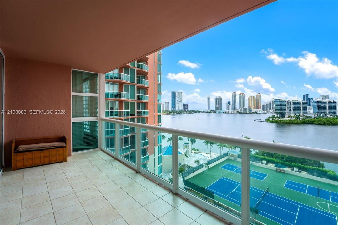 Covered balcony featuring tile flooring and a glass-paneled railing overlooking a tennis court and waterfront cityscape