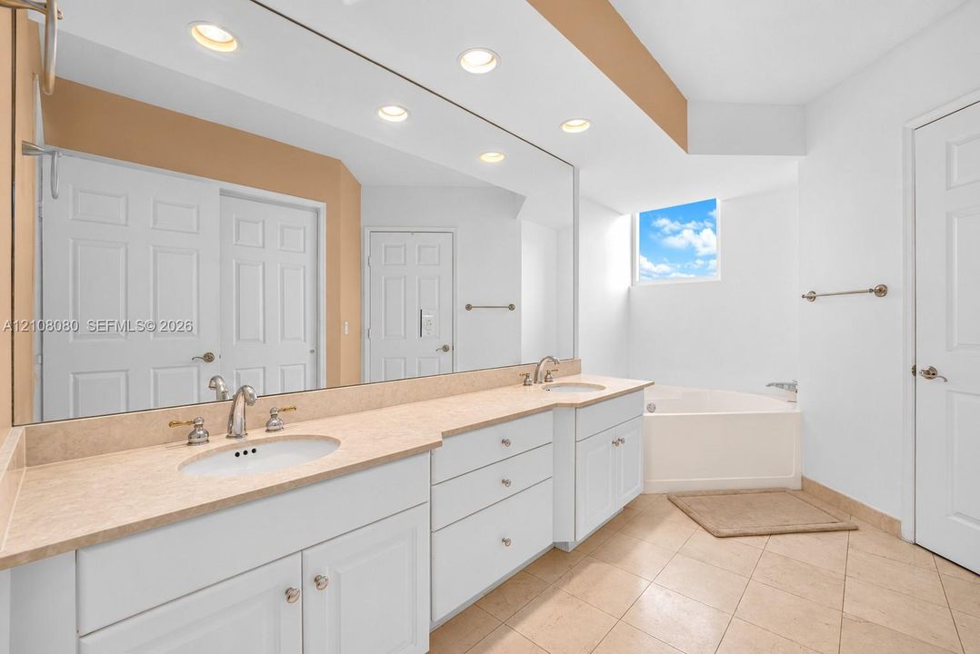 Dual-sink vanity with neutral countertop, expansive wall-to-wall mirror, recessed lighting, tile flooring, and built-in soaking tub