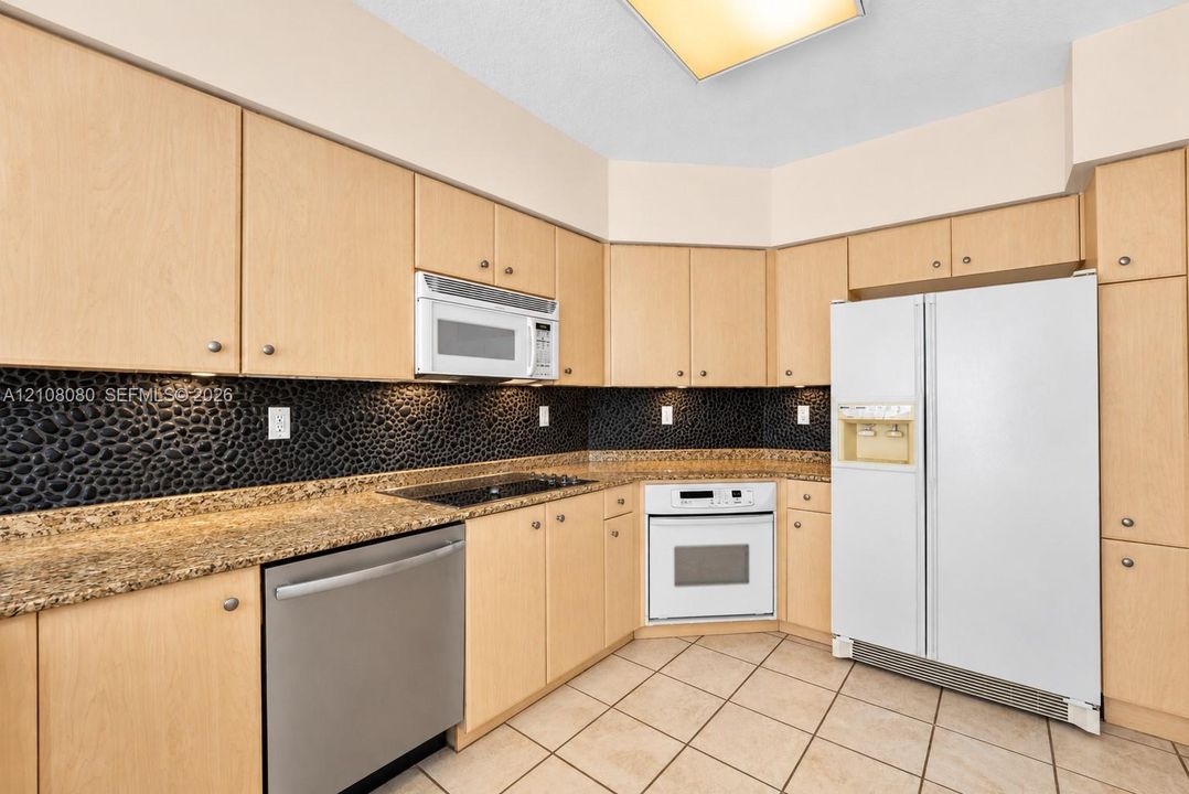 Kitchen featuring light wood-finish cabinetry, stone-texture countertops, black pebble-style backsplash, tile flooring, and built-in appliances