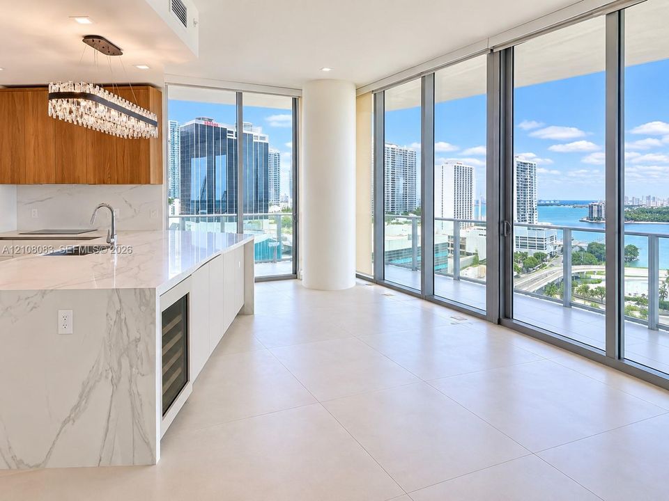Open-concept kitchen featuring an expansive waterfall-edge island, stone-surfaced countertops, custom wood cabinetry, integrated wine refrigerator, and floor-to-ceiling glass windows