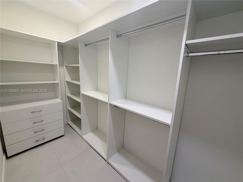 Custom walk-in closet featuring white built-in shelving, multiple hanging rods, integrated four-drawer dresser, and tile flooring