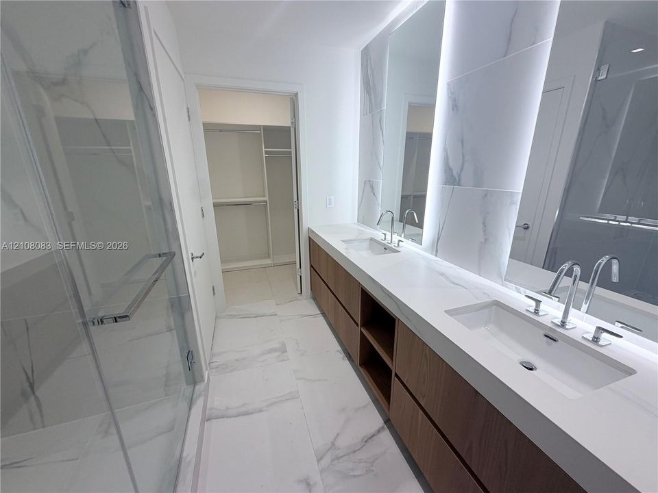 Dual-vanity bathroom featuring a floating wood-finish cabinet with integrated shelving, quartz countertops, floor-to-ceiling stone-style tile flooring, and a glass-enclosed shower