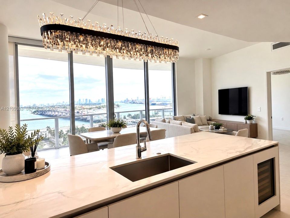 Kitchen island featuring stone-finish countertops, an undermount rectangular sink, and a built-in wine refrigerator, accented by a linear crystal chandelier and floor-to-ceiling windows