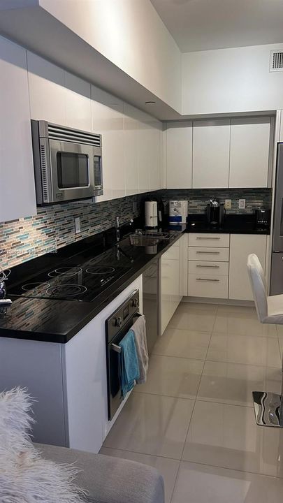 Kitchen featuring high-gloss white cabinetry, polished dark stone countertops, linear glass mosaic tile backsplash, integrated stainless steel appliances, and large-format tile flooring