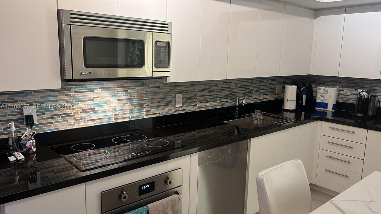 Kitchen featuring white slab cabinetry, black countertops, multi-toned glass tile backsplash, stainless steel built-in microwave, and electric cooktop