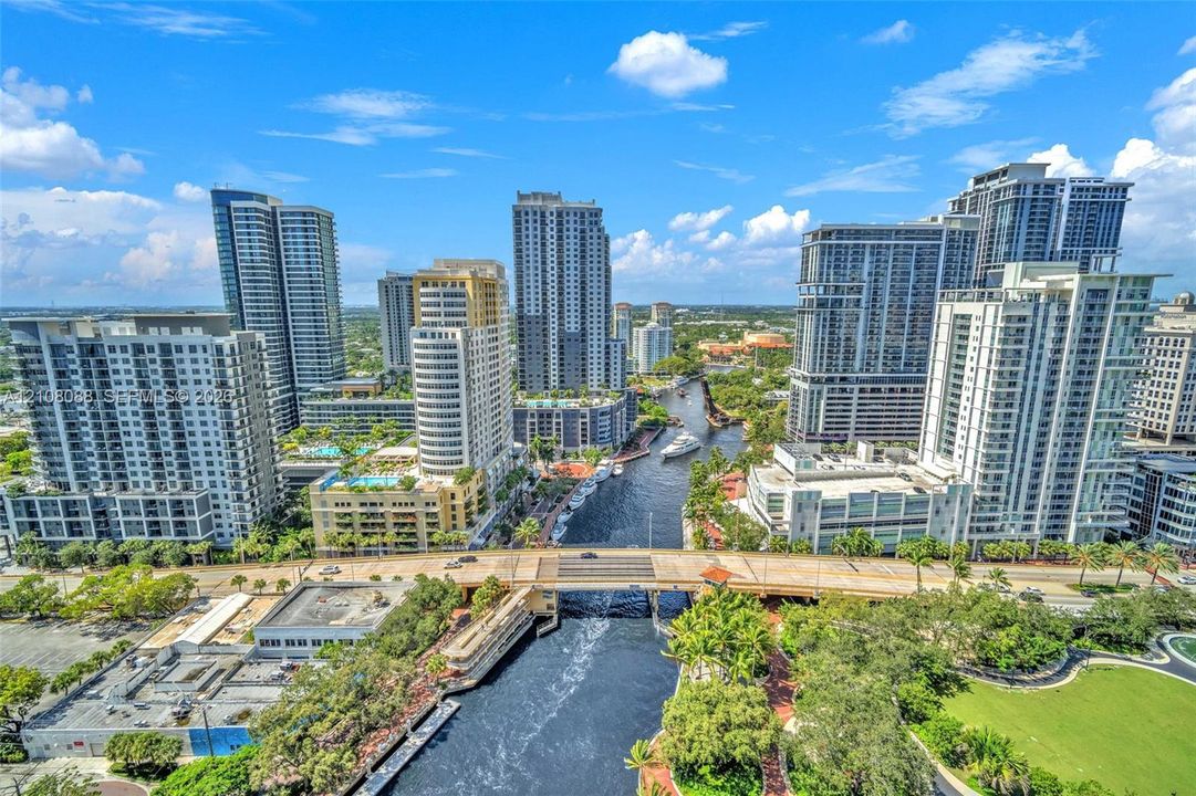 Aerial perspective of a high-density urban corridor featuring a central waterway, bridge infrastructure, mature landscaping, and numerous mid-rise and high-rise structures