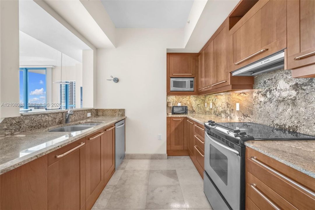 Kitchen featuring wood cabinetry, stone countertops, stone backsplash, stainless steel appliances, and large-format tile flooring