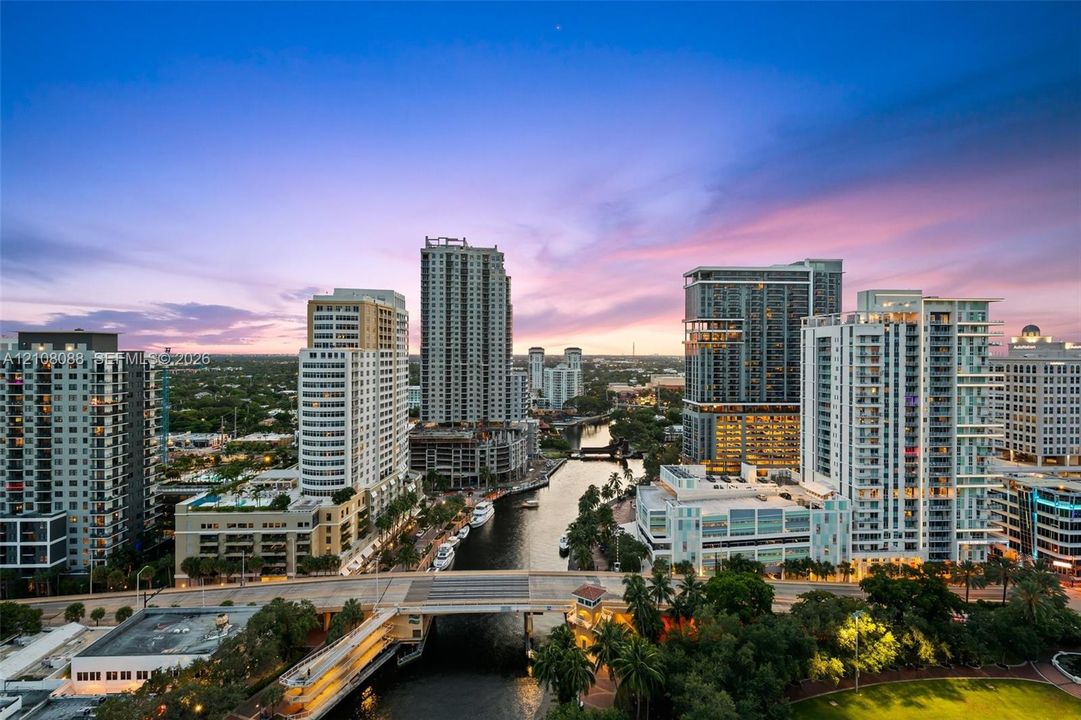 Aerial perspective featuring a metropolitan skyline, a central waterway, bridge infrastructure, and dense urban development