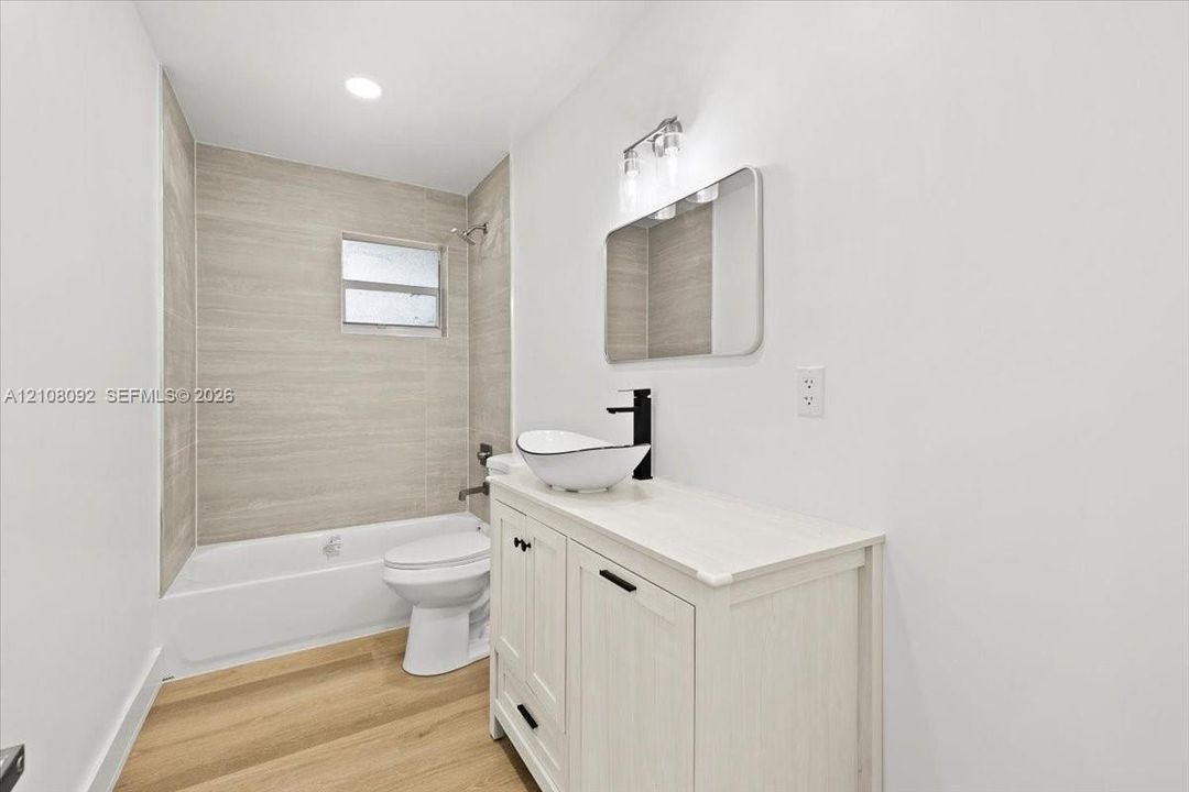 Bathroom featuring wood-finish flooring, a white vanity with a vessel sink, a matte black faucet, and a tiled tub surround