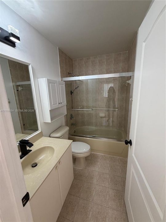 Bathroom featuring a solid-surface vanity countertop, matte black faucet, wall-mounted white cabinetry, tile flooring, and a glass-enclosed tub and shower combination with tiled surround