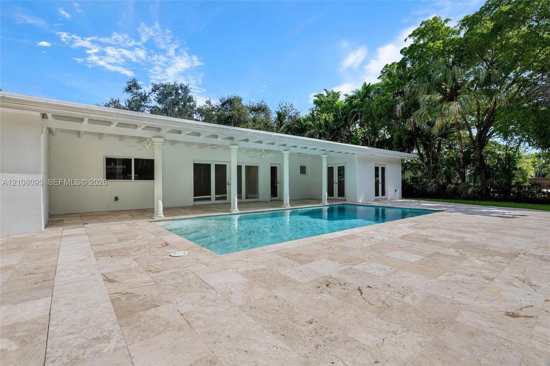 In-ground swimming pool featuring a sprawling stone patio, covered lanai supported by classical columns, and multi-panel glass door entries