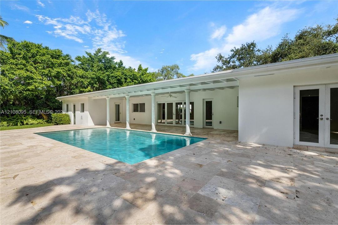 Rectangular in-ground pool with stone paver decking, covered patio supported by classical columns, and multi-panel glass entry doors