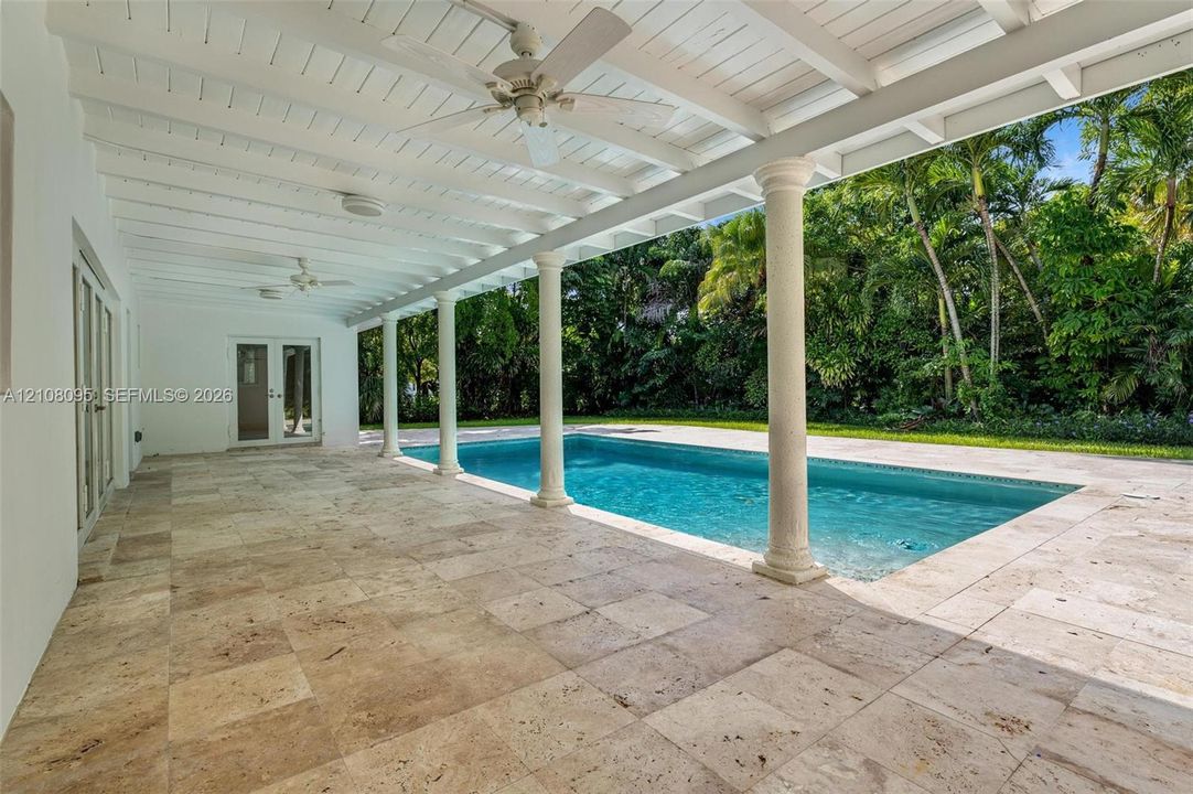 Covered patio featuring stone-tiled flooring, white beamed ceilings, and ceiling fans, opening onto a swimming pool with structural columns and perimeter greenery