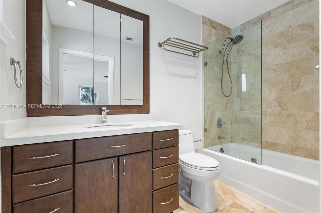 Dark-finish wood cabinetry, integrated rectangular sink with stone-surface countertop, full-width framed mirror, bathtub with glass panel partition, and stone-tile shower surround