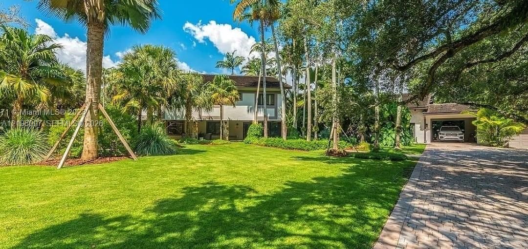 Two-story residence featuring a multi-pitched roof, paved stone driveway, attached garage, and expansive lawn shaded by mature palms and canopy trees