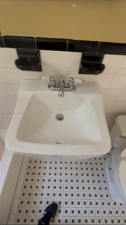 Wall-mounted porcelain sink with chrome dual-handle faucet, built-in ceramic soap and toothbrush holders, and black-and-white geometric tile flooring