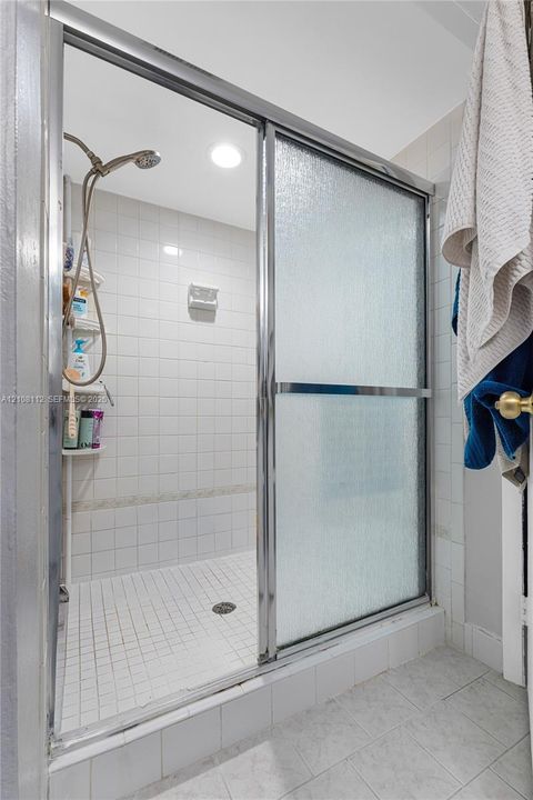 Shower enclosure featuring a chrome-framed sliding glass door, white tiled walls, a handheld shower fixture, and a built-in corner shelving unit