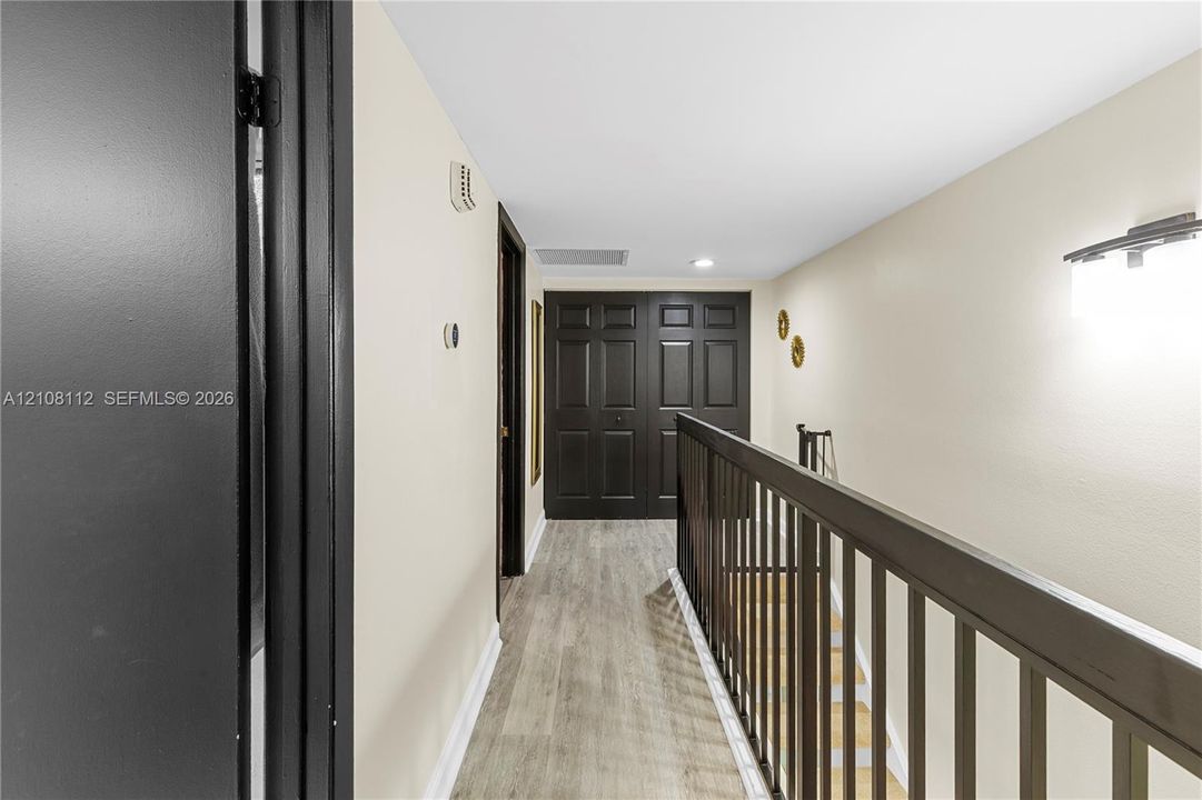 Hallway featuring wood-finish flooring, dark-toned interior doors, and a matching handrail with vertical balusters