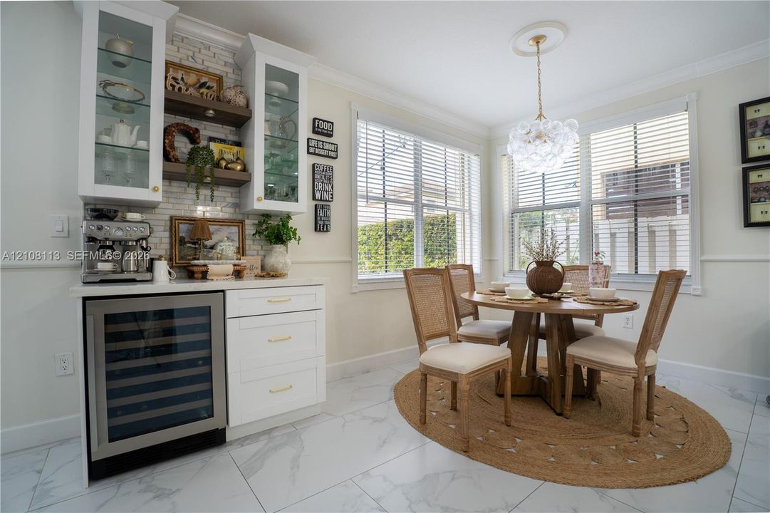 Breakfast area featuring white cabinetry with glass-front uppers, built-in wine refrigerator, white subway tile backsplash, crown molding, and large windows with horizontal blinds