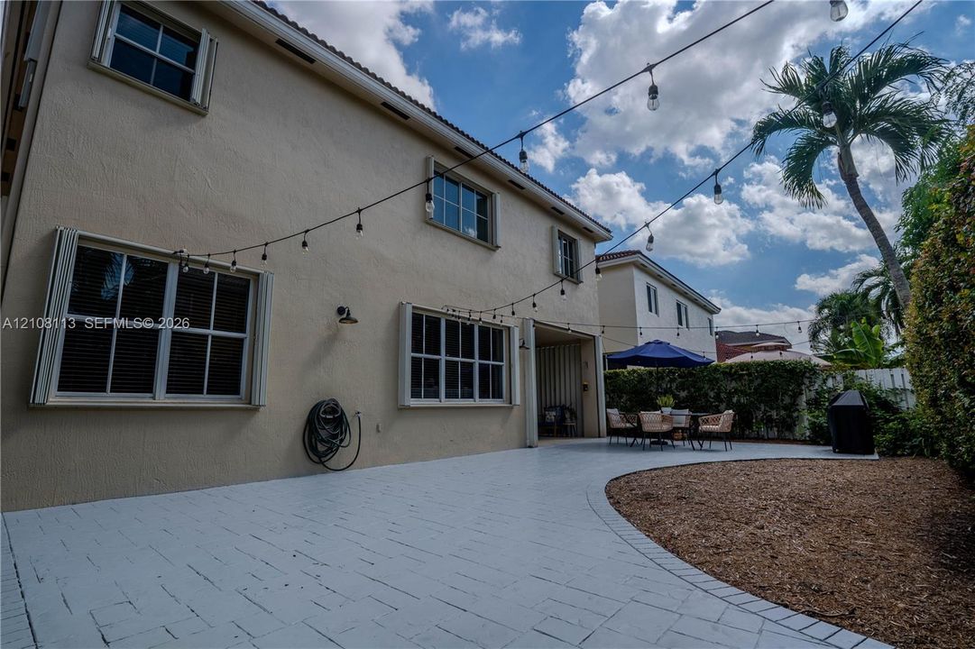 Expansive paved patio featuring integrated string lighting, stucco exterior cladding, multi-pane windows, and manicured landscaping