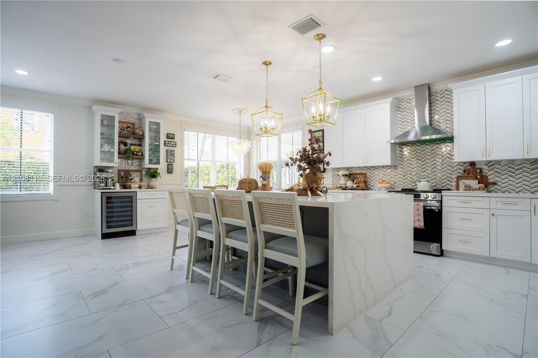 Kitchen featuring a central island with stone-finish waterfall edges, herringbone-patterned tile backsplash, white shaker-style cabinetry, stainless steel range hood, and porcelain tile flooring