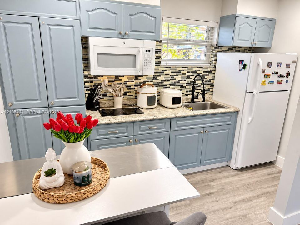 Kitchen featuring blue cabinetry, light-toned stone-finish countertops, decorative multi-tone tile backsplash, stainless steel sink with a high-arch matte black faucet, and wood-finish flooring