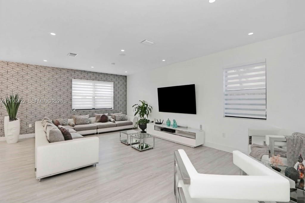 Living area featuring light-toned wood-finish flooring, recessed lighting, geometric-patterned accent wall, and dual-layered window shades