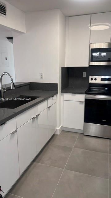 Kitchen featuring high-gloss white cabinetry, dark stone-finish countertops, stainless steel appliances, a pull-down kitchen faucet, and large-format tile flooring