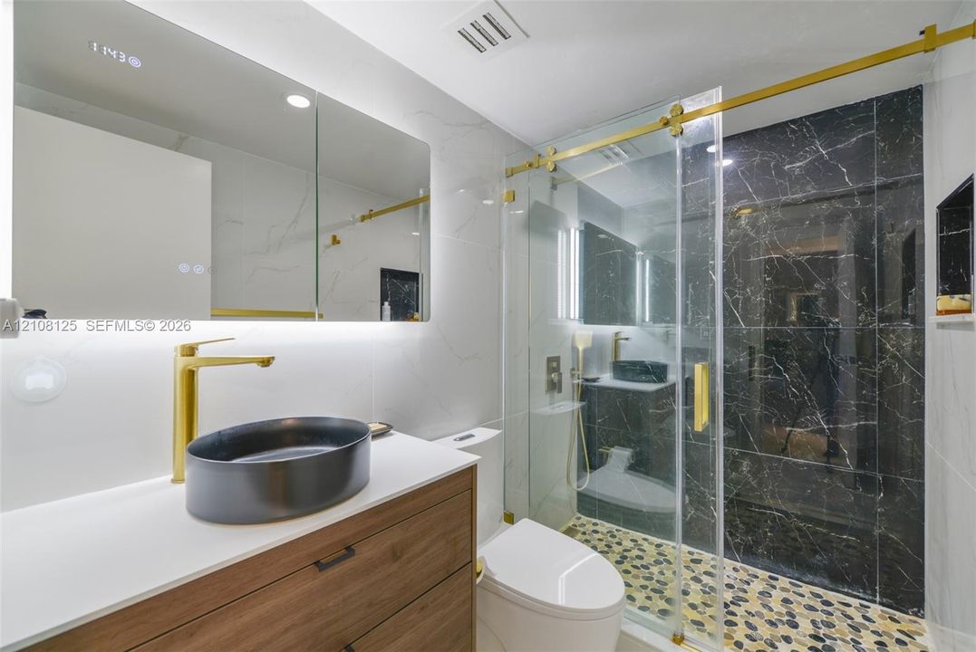 Bathroom featuring a wood-finish vanity with a quartz countertop, oval vessel sink, gold-toned hardware, and a walk-in shower with stone-tile flooring and dark accent tiling