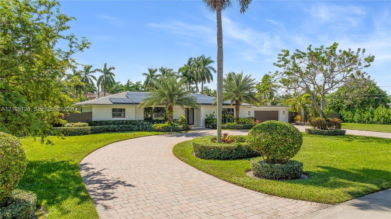 Single-story exterior featuring a paver circular driveway, solar panel installation, manicured landscaping with mature palm trees, and an attached two-car garage