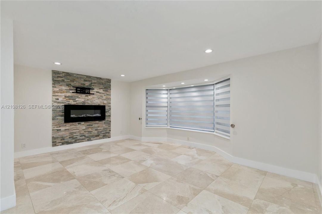 Stone-accented fireplace with a wall-mounted fire feature, large bay window, and light-toned tile flooring