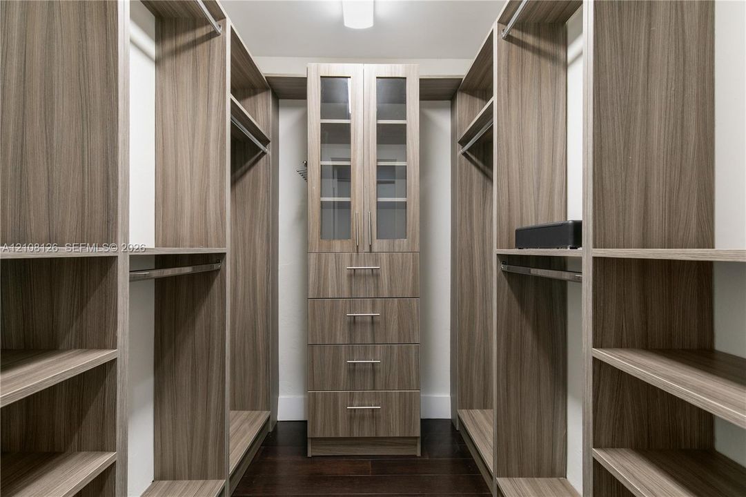 Custom walk-in closet featuring extensive wood-finish shelving, integrated hanging rods, a central stack of four soft-close drawers, and upper cabinetry with glass-panel doors