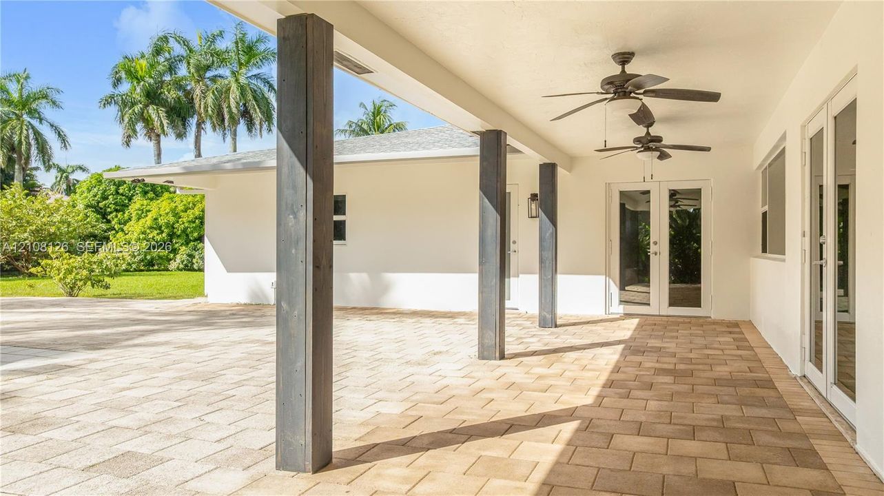 Covered outdoor patio featuring light-toned paver flooring, dark wood-finish structural support columns, dual ceiling fans, and double glass-paned doors