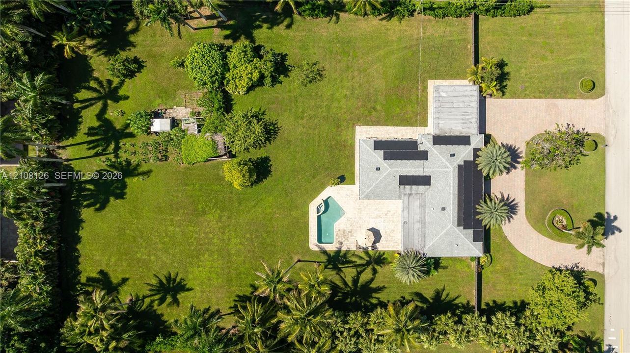 Aerial perspective featuring an expansive landscaped lot, paved circular driveway, integrated swimming pool with stone-surfaced patio, and a shingled roof with solar panels