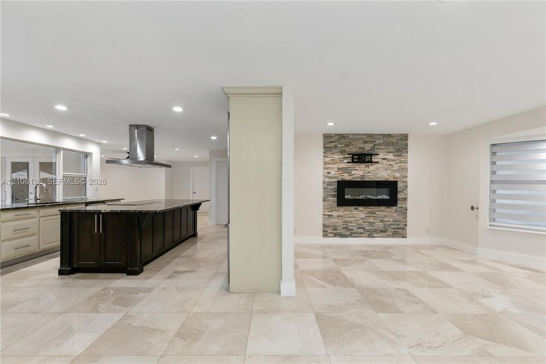 Kitchen featuring a dark-finish island with a countertop, stainless steel range hood, stone-finish fireplace, and neutral-tone tile flooring