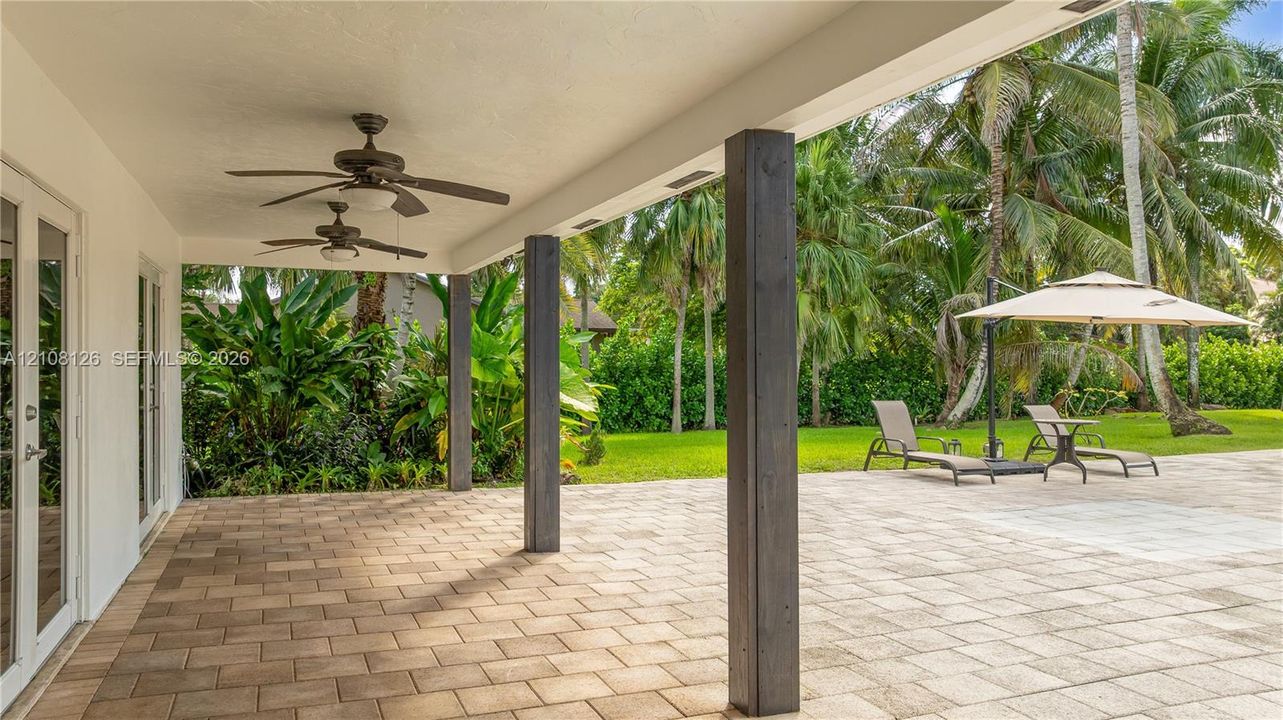 Covered patio featuring concrete paver flooring, dual ceiling fans, and dark-stained structural support pillars